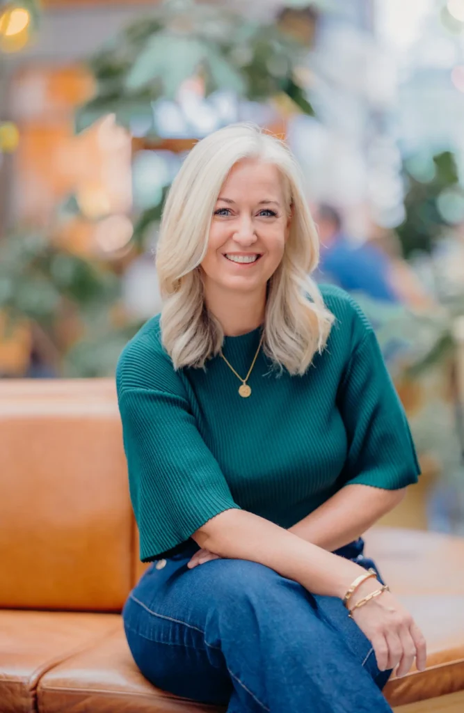 Erica Wood smiling warmly while seated indoors, representing a friendly and approachable media training expert.